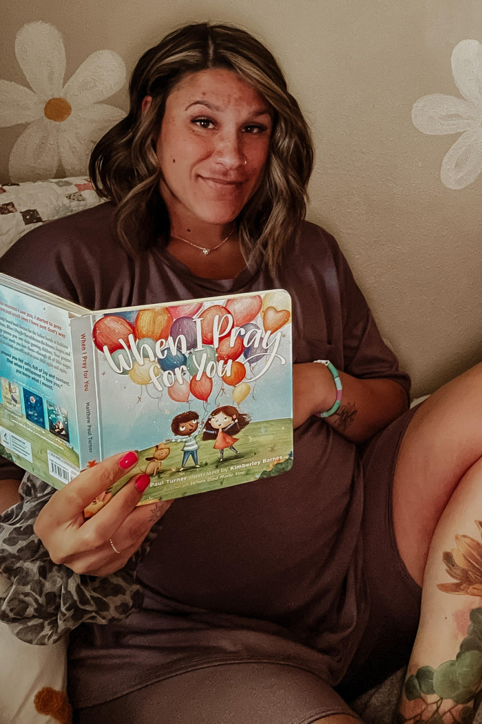woman wearing a gray maternity lounge set and holding a children's book while sitting on a bed with a visible baby bump