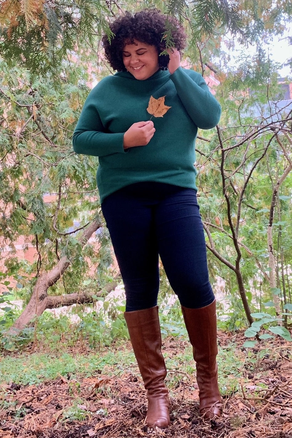 woman wearing a green sweater with a leaf detail and dark jeans, standing outdoors in tall brown boots