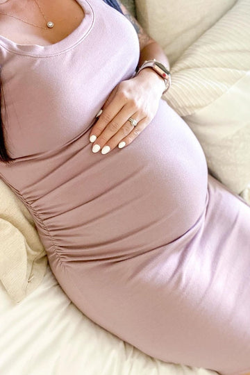 woman wearing a fitted mauve maternity dress with side ruching, gently holding her baby bump while sitting on a bed