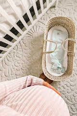woman wearing a light pink striped maternity dress with a visible baby bump, beside a woven baby basket in a nursery setting