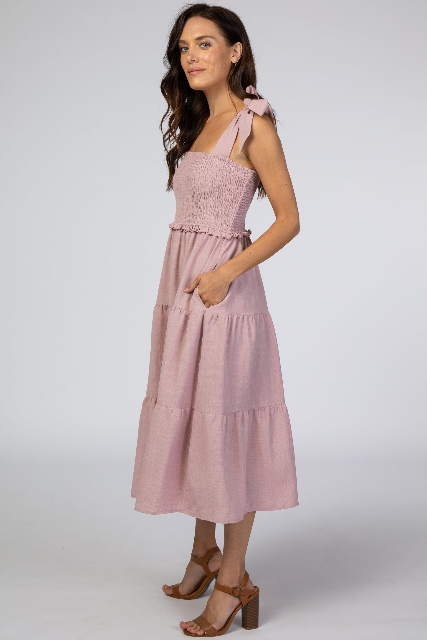 woman wearing a light pink smocked sundress with tie straps and tiered skirt, standing profile with hands in pockets