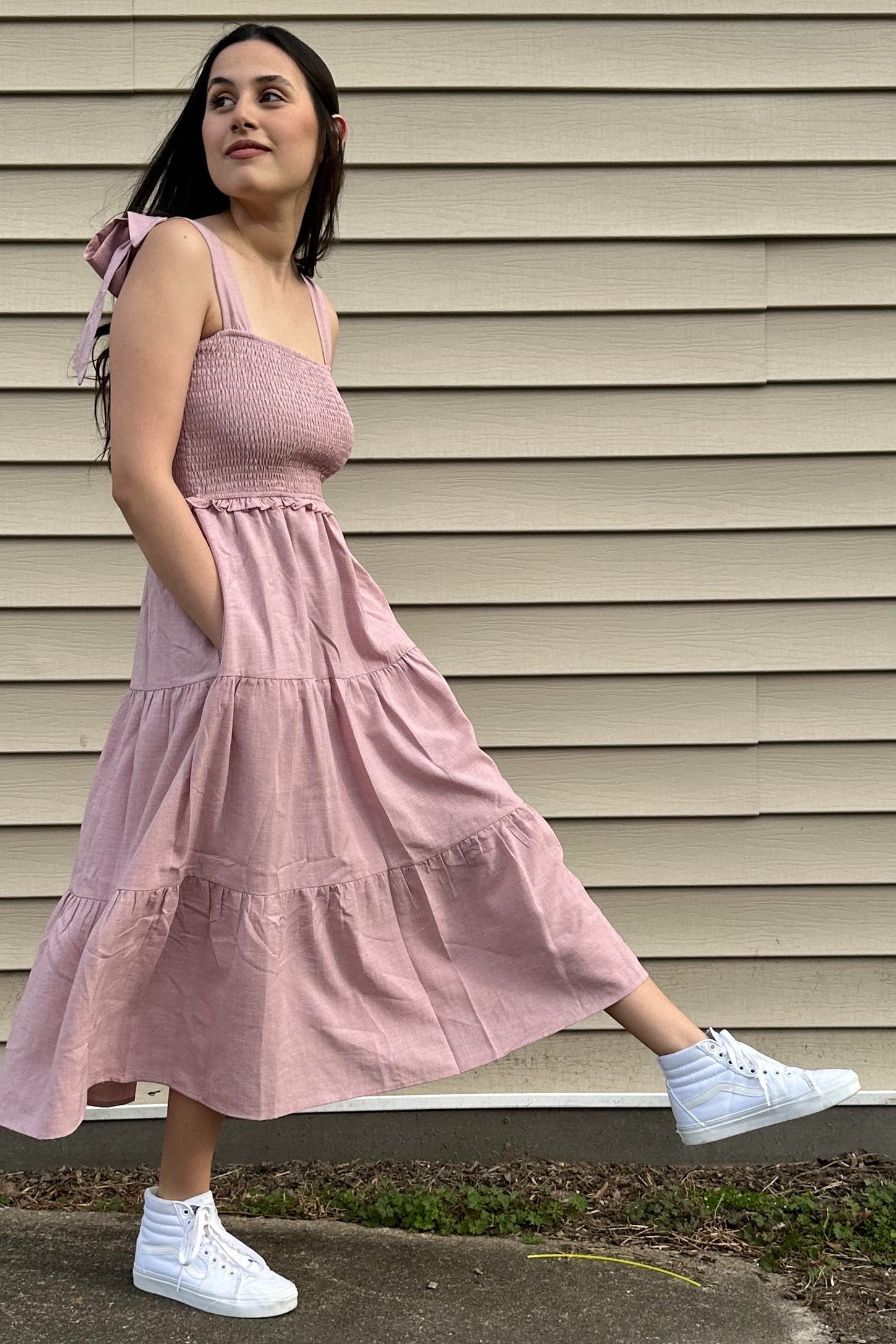 woman wearing a pink smocked maxi dress with tie straps and pockets, styled with white sneakers.