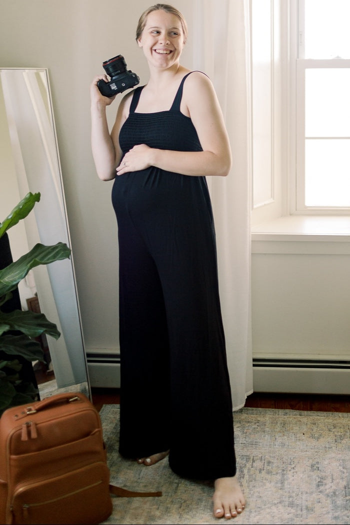 woman wearing a black maternity jumpsuit holding a camera and showing her baby bump
