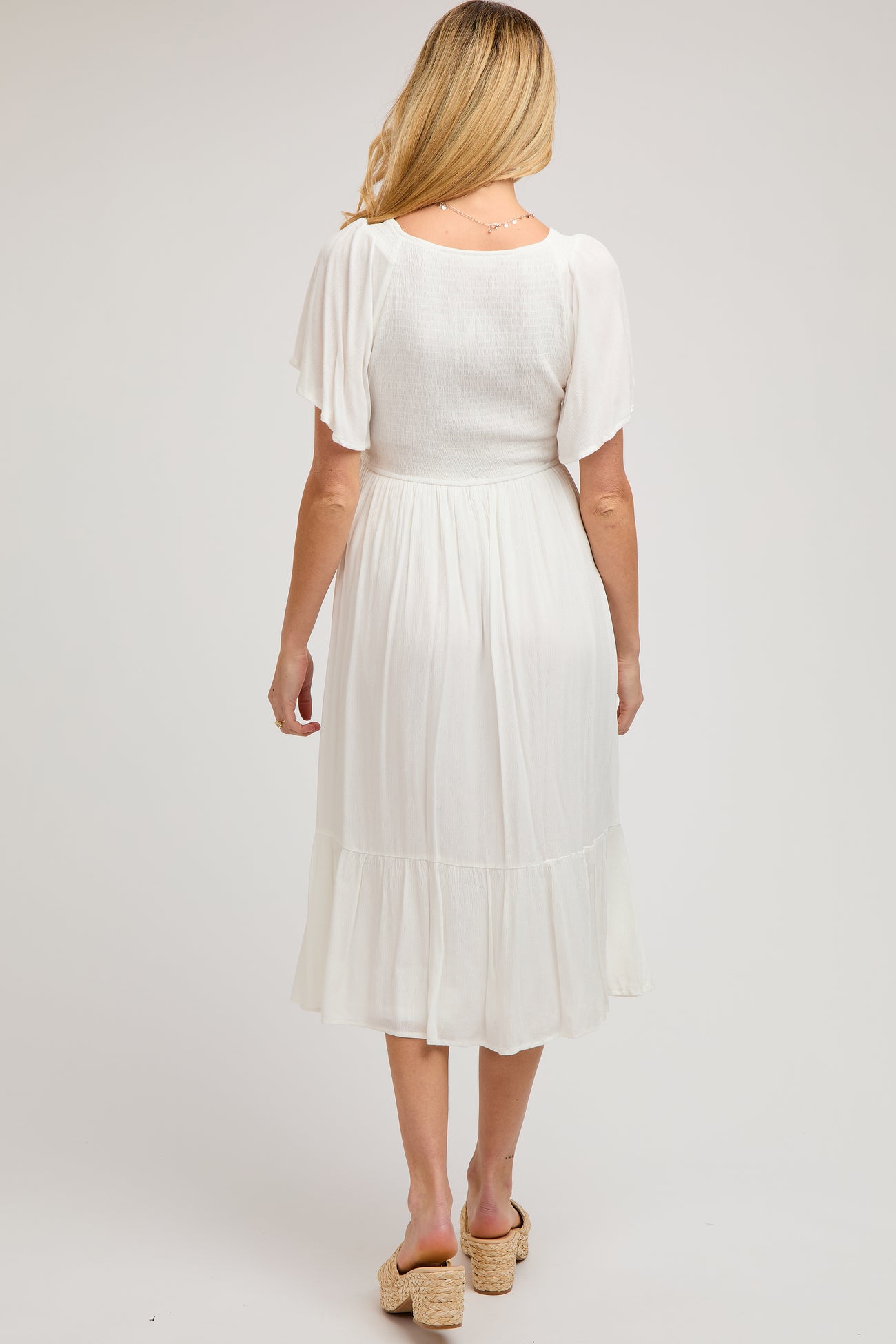 woman wearing a white maternity maxi dress with short flutter sleeves and a tiered ruffle hem, showing her back