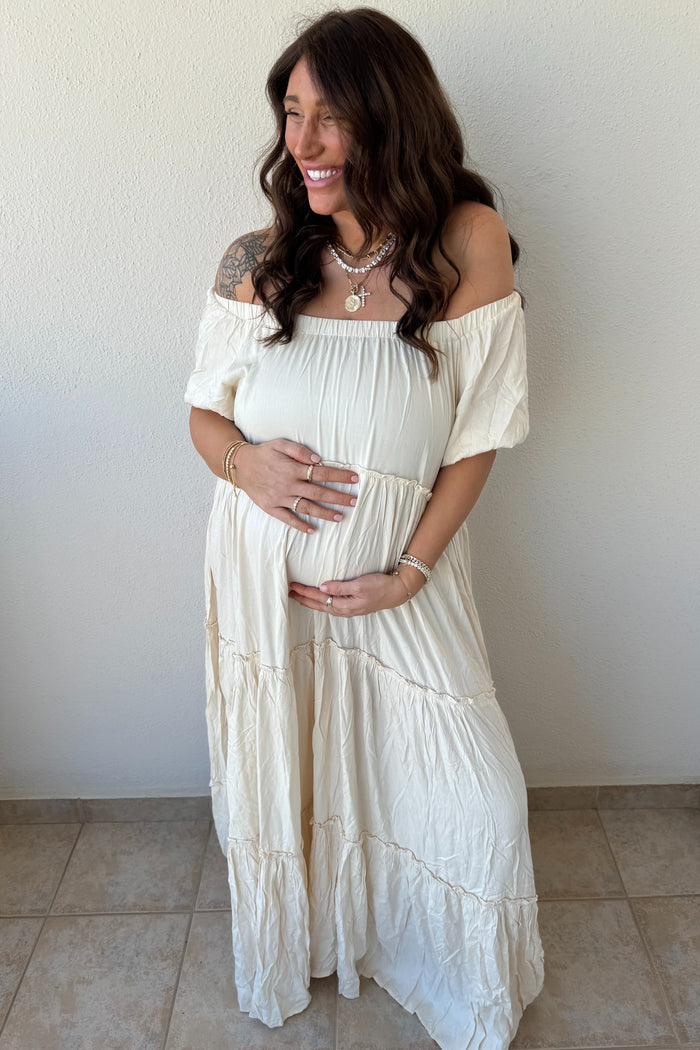 woman wearing a white off-shoulder maternity maxi dress and holding her baby bump