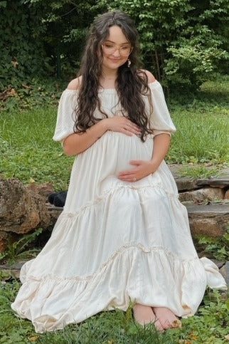 woman wearing a cream off-the-shoulder maternity dress and holding her baby bump outdoors