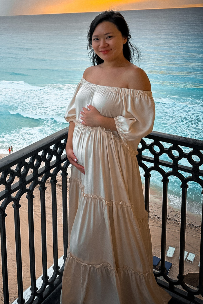 woman wearing a cream off-shoulder maternity dress with tiered details and a visible baby bump standing by the ocean