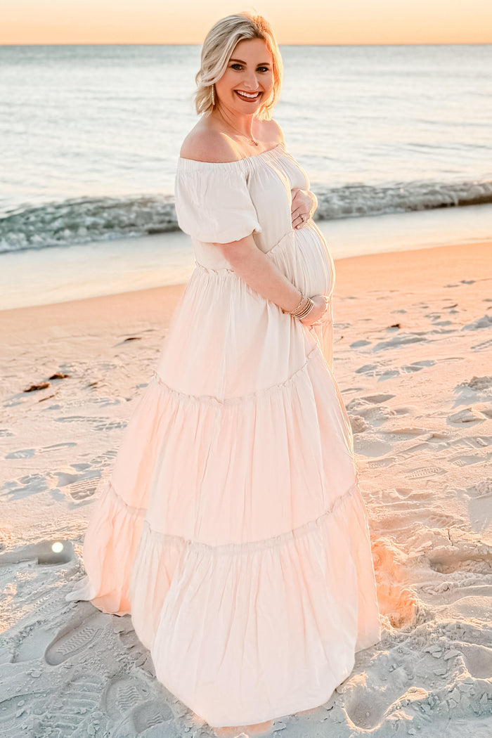 woman wearing a blush pink off-shoulder maternity gown with tiered layers, cradling her baby bump on the beach at sunset