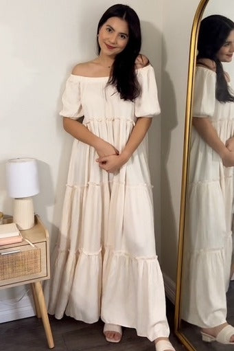 woman wearing a white off-shoulder maxi dress with tiered layers, standing beside a mirror and smiling