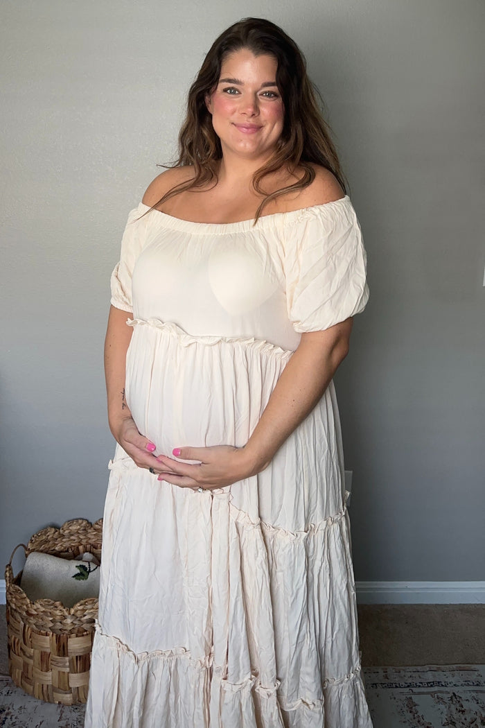 woman wearing a soft peach off-shoulder maternity dress with flowing layers and holding her baby bump