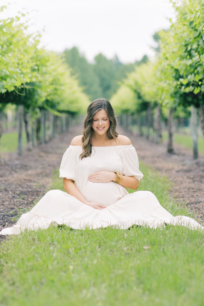 woman wearing a white off-the-shoulder maternity dress sitting on the grass and holding her baby bump in a vineyard setting