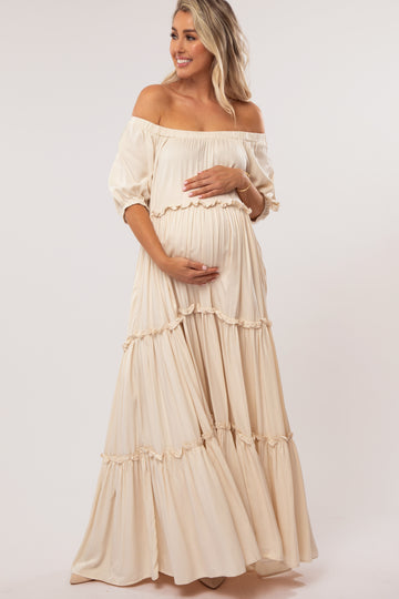 woman wearing a cream off-the-shoulder maternity maxi dress with ruffle details and cradling her baby bump