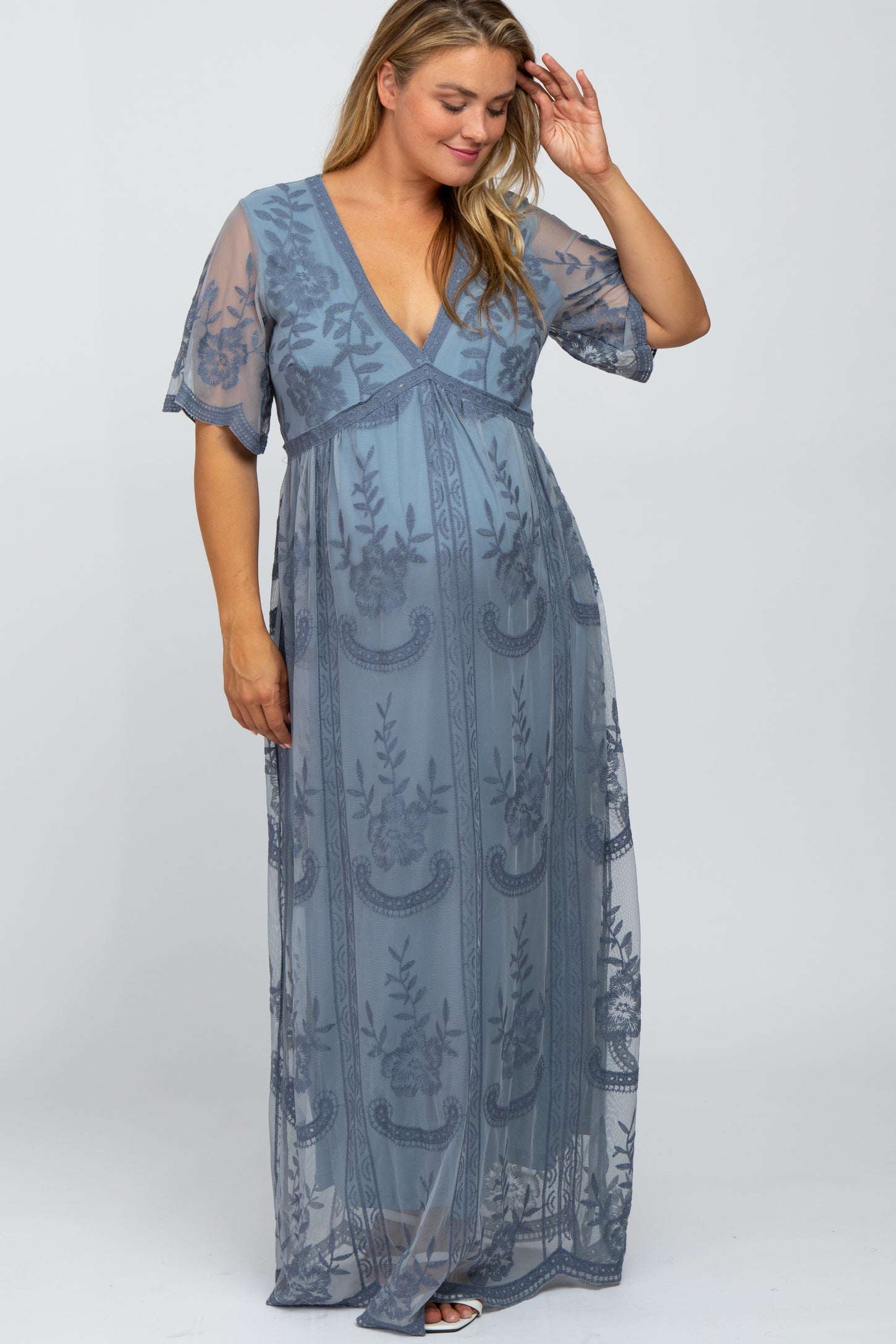 woman wearing a blue maternity plus sheer maxi dress with floral embroidery and short sleeves, showing her baby bump