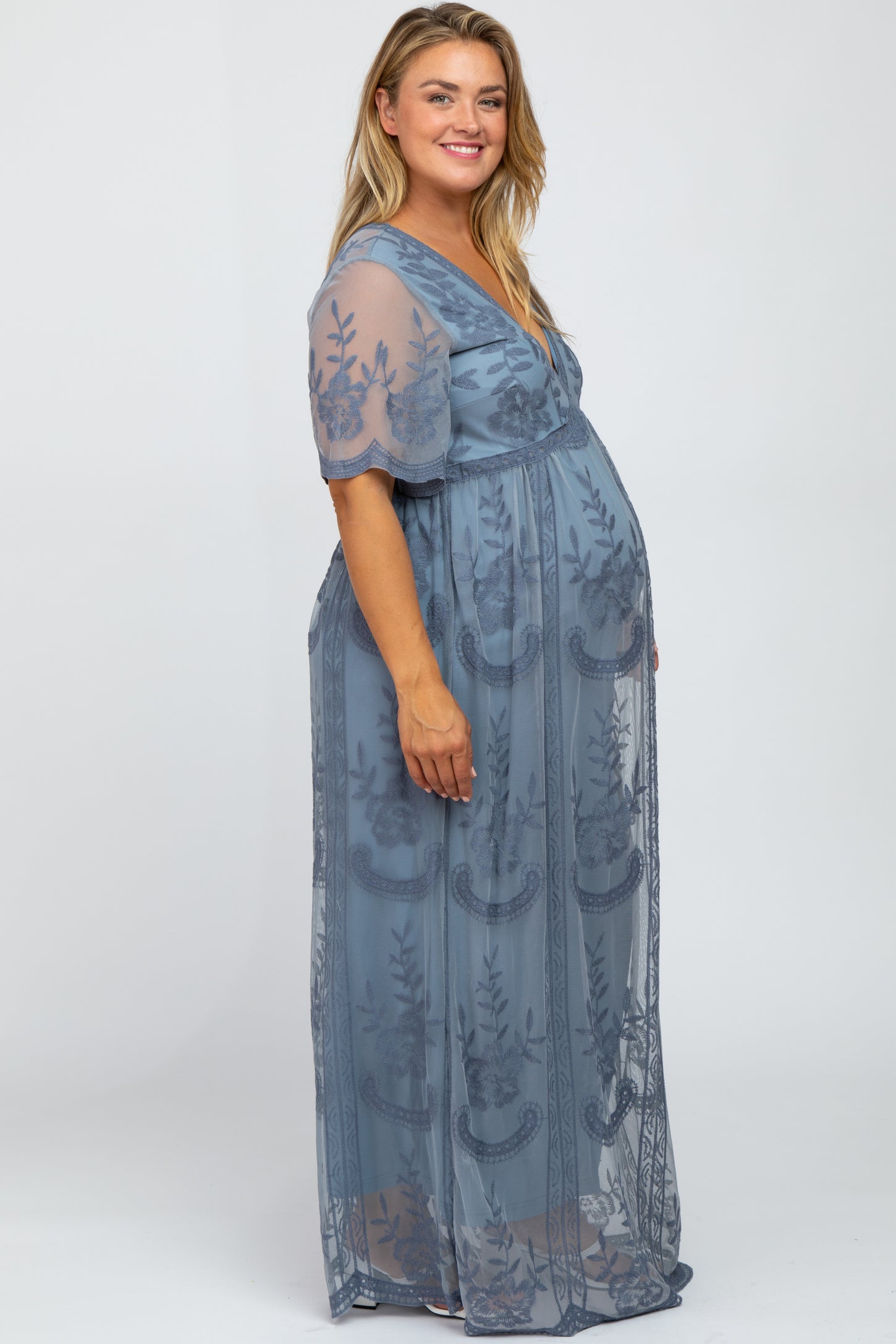 woman wearing a sheer blue maternity plus gown with floral embroidery and a visible baby bump