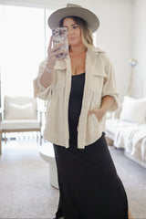 woman wearing a beige corduroy shirt jacket over a black maxi dress, holding her phone and posing in a bright living room