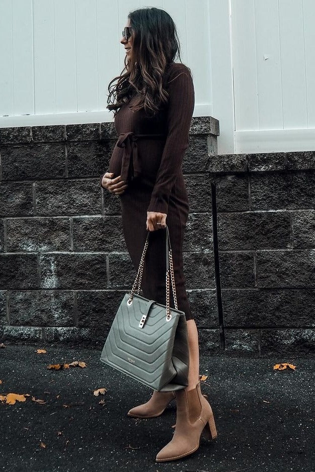 woman wearing a brown maternity dress with a tie waist and a visible baby bump, carrying a gray handbag and wearing ankle boots