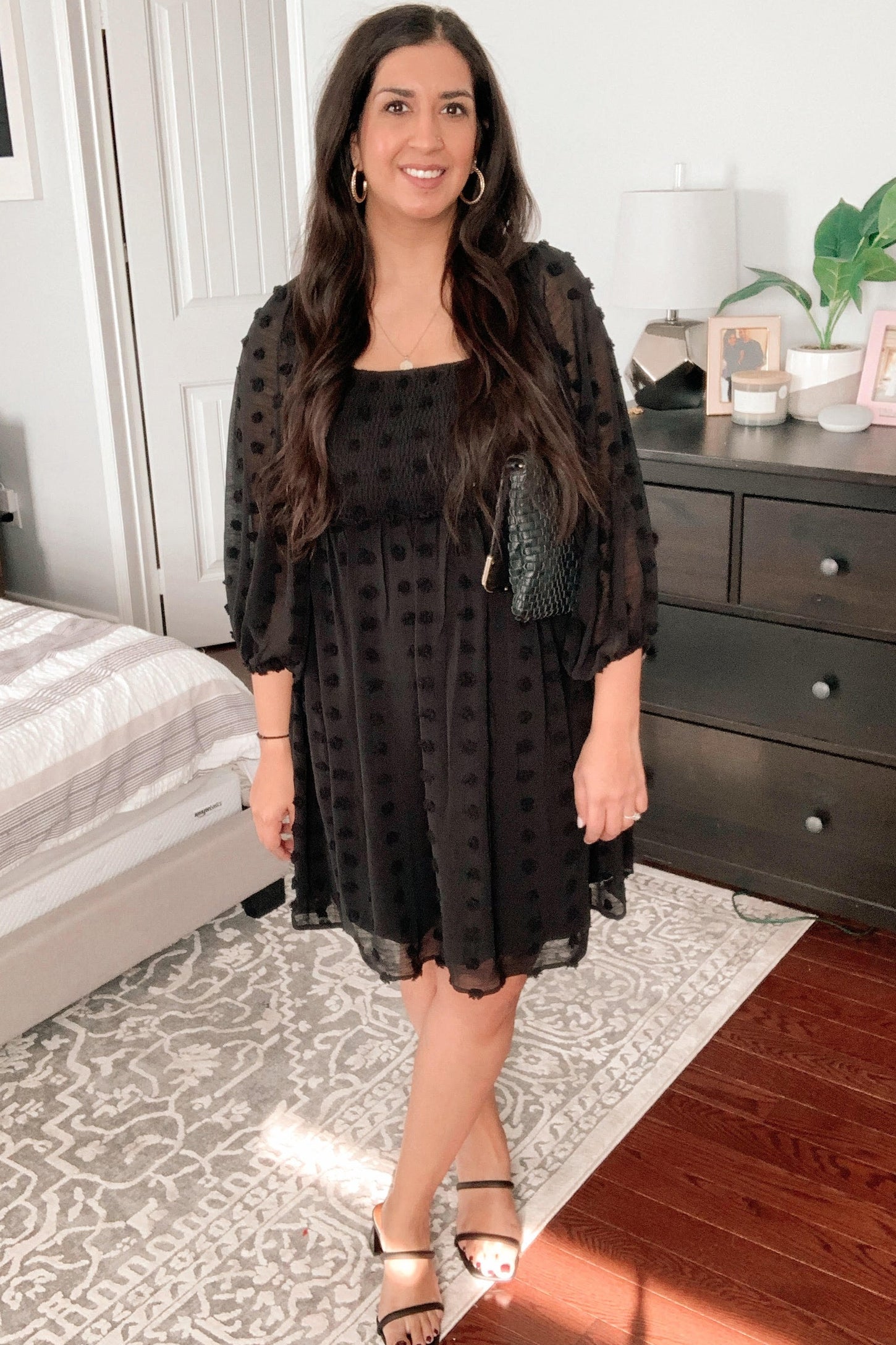 woman wearing a black puff sleeve dress with textured dots and sandals, standing in a bedroom setting