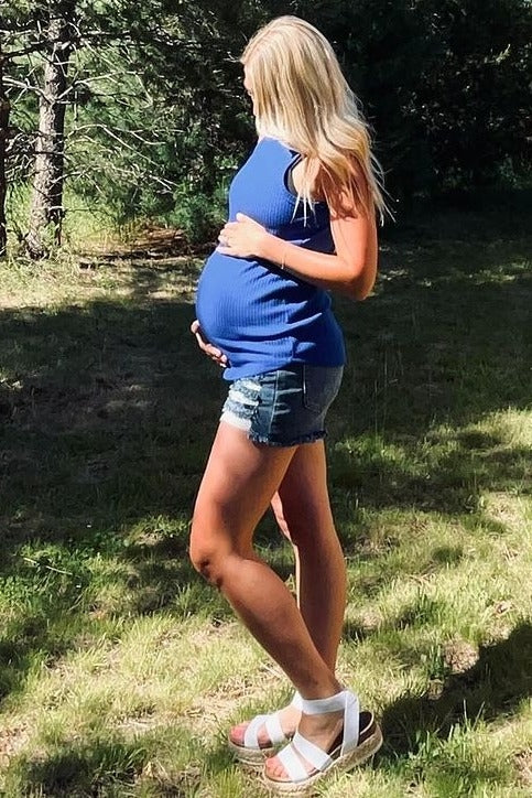 woman wearing a blue ribbed maternity tank top and denim shorts, cradling her baby bump in a sunny outdoor setting