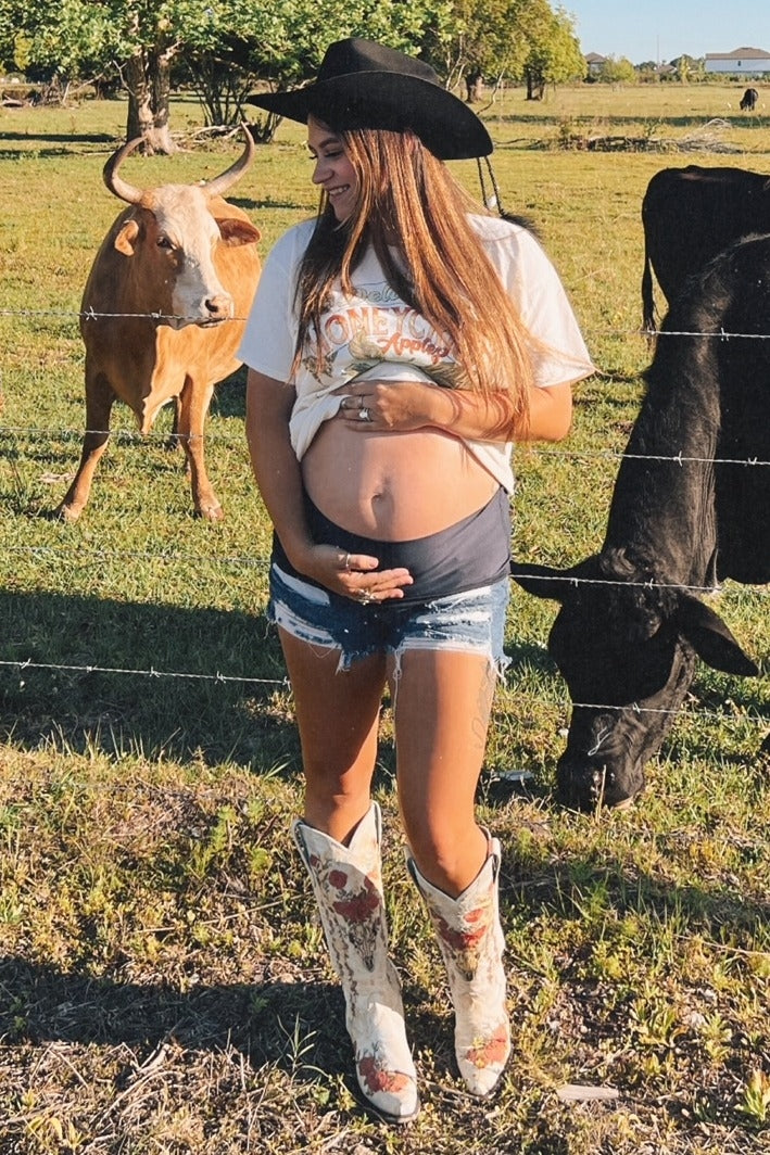 woman wearing a white graphic t-shirt and denim shorts, showing baby bump, with cowboy boots in a field with cows