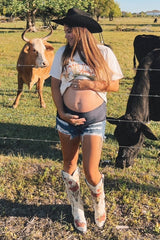 woman wearing a white graphic t-shirt and denim shorts, showing baby bump, with cowboy boots in a field with cows