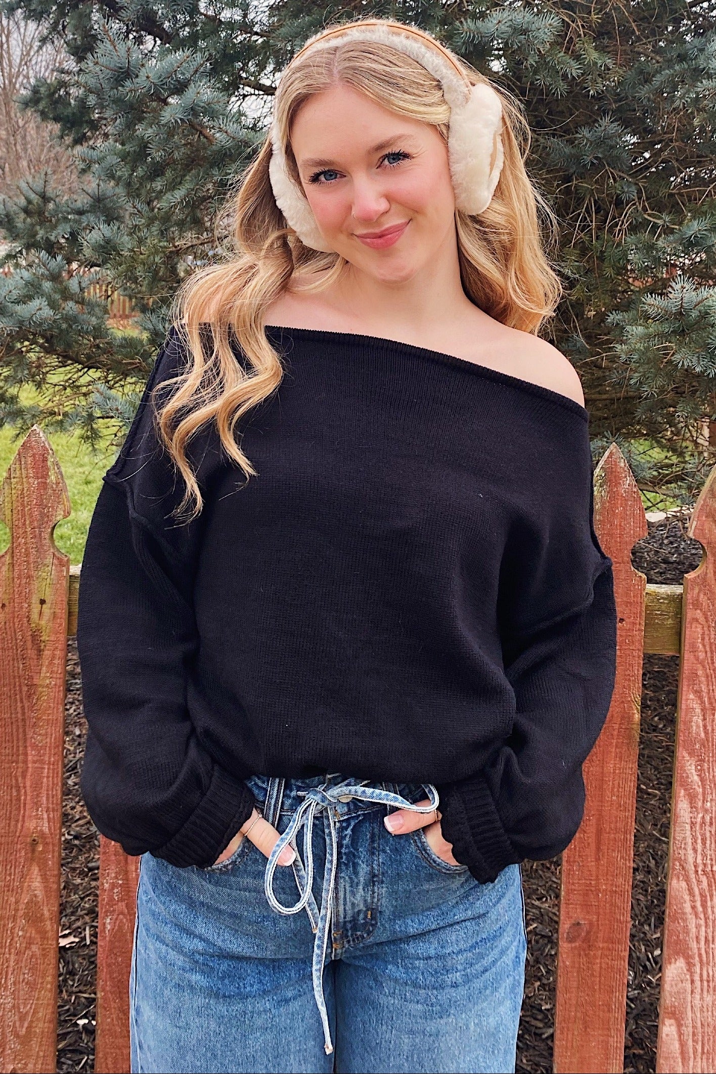 woman wearing a black off-the-shoulder sweater and light blue high-waisted jeans, standing near a wooden fence