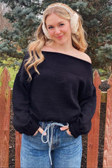 woman wearing a black off-the-shoulder sweater and light blue high-waisted jeans, standing near a wooden fence