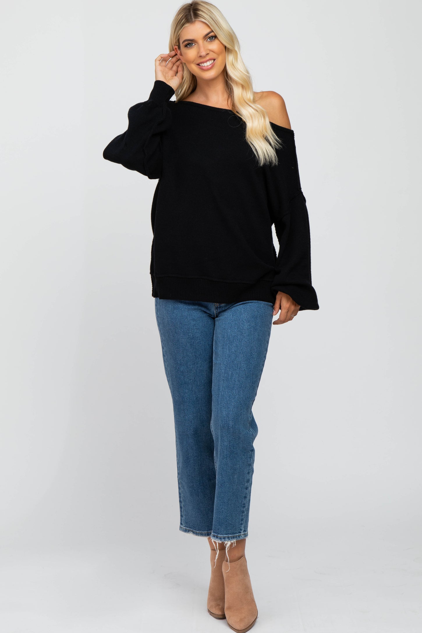 woman wearing a black off-the-shoulder sweater with bell sleeves and blue high-waisted jeans, standing with one hand on her hair