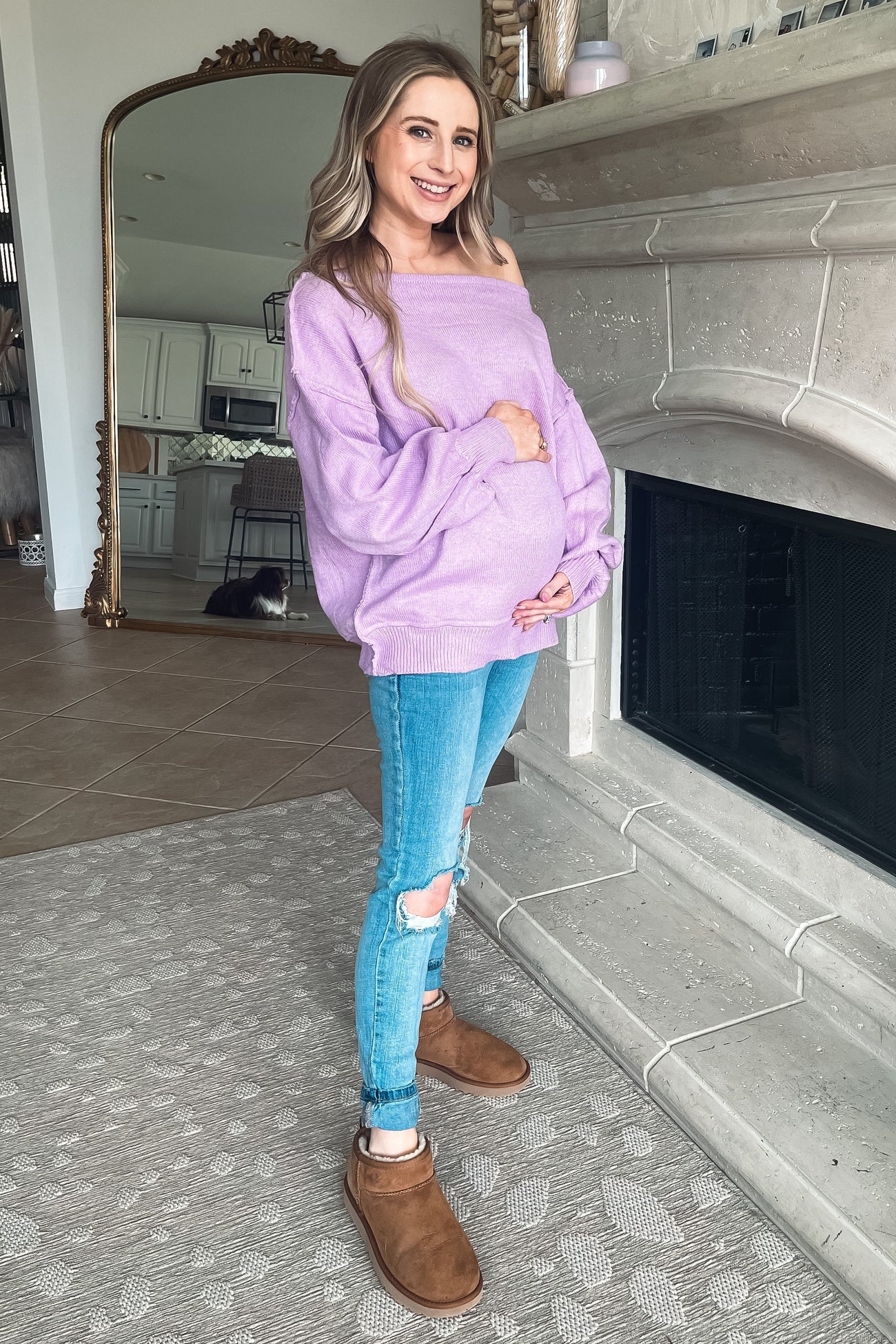 pregnant woman in purple off-the-shoulder sweater and ripped jeans standing by a fireplace
