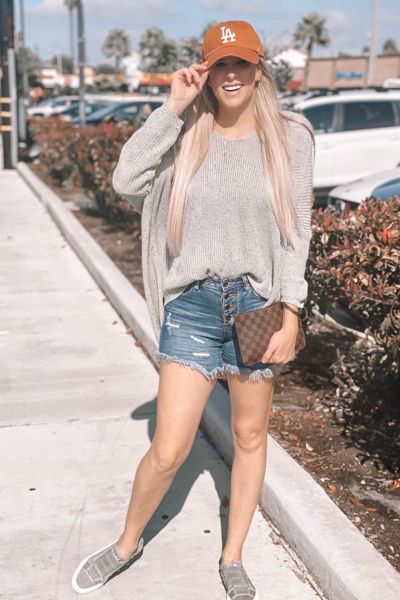 woman wearing a light gray oversized sweater and distressed denim shorts, holding a brown handbag and wearing a baseball cap