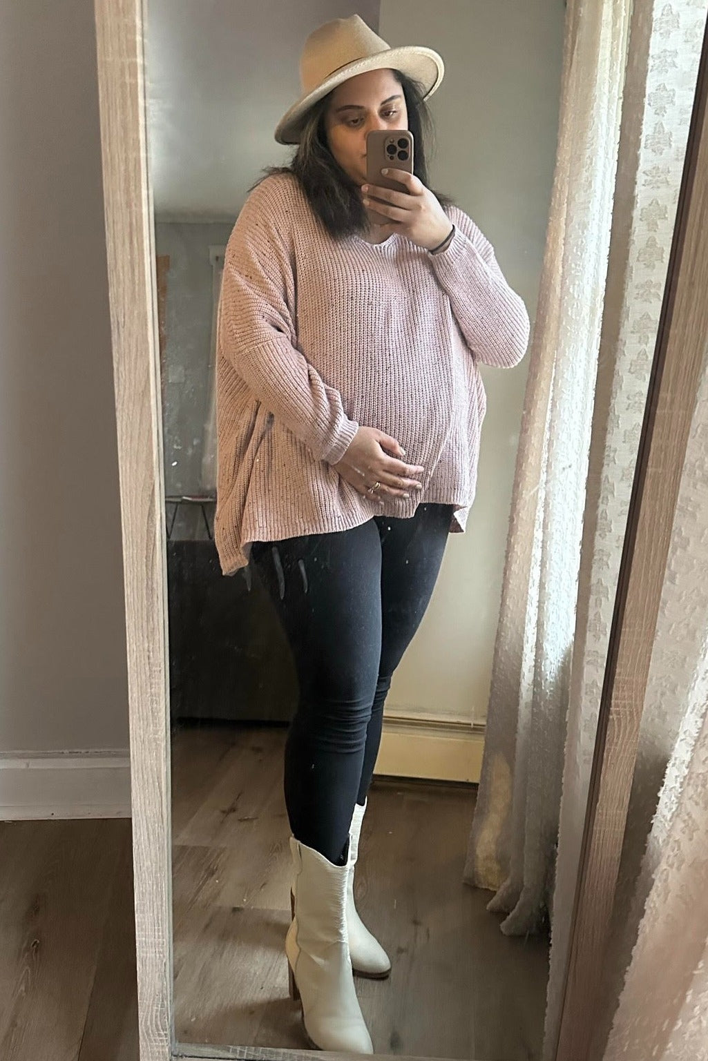 woman wearing a light pink oversized maternity sweater, black leggings, and white boots, holding her baby bump in front of a mirror