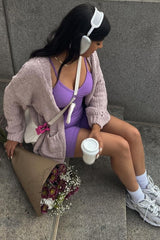 woman wearing a light purple fitted dress and a chunky pink cardigan, holding a coffee cup and flowers, with headphones on