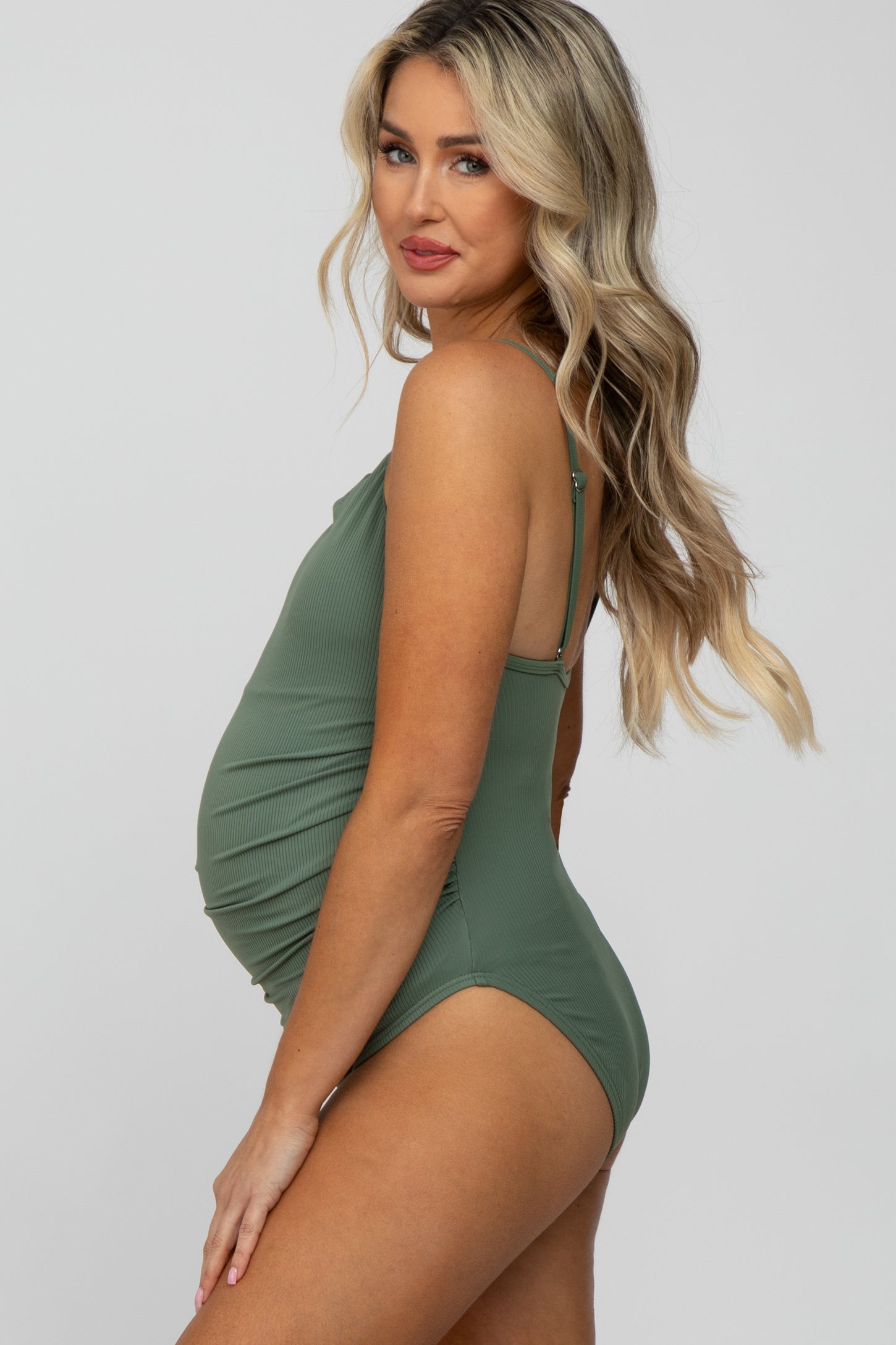 woman wearing a sage green maternity swimsuit with adjustable straps and ruching, showcasing her baby bump