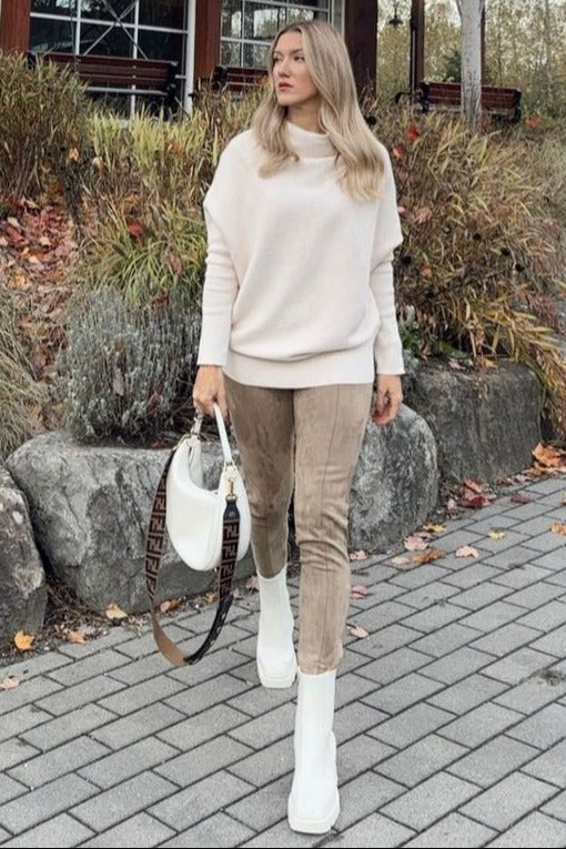woman wearing a light pink oversized sweater and beige suede pants, carrying a white handbag, walking outdoors