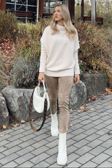 woman wearing a light pink oversized sweater and beige suede pants, carrying a white handbag, walking outdoors