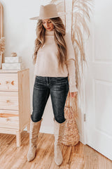 woman wearing a beige sweater, dark jeans, and beige knee-high boots, styled with a wide-brim hat and posing indoors
