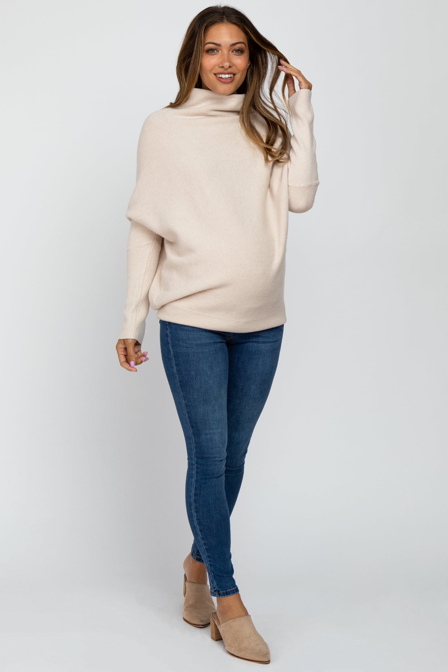 woman wearing a light beige maternity sweater with a draped neckline and blue skinny jeans, showing her baby bump
