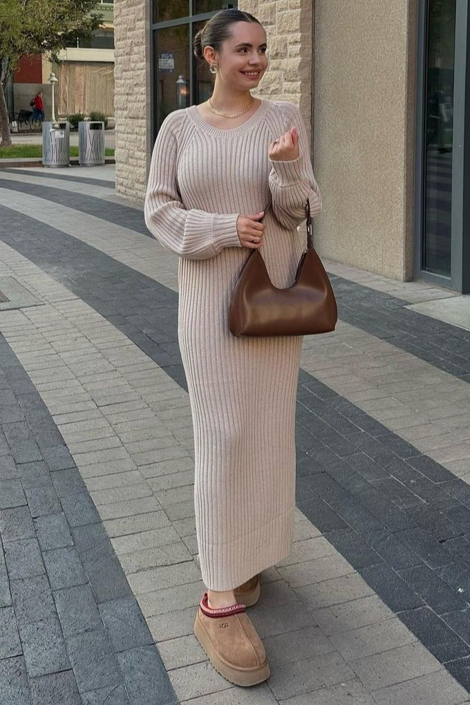 woman wearing a beige ribbed knit sweater dress, holding a brown handbag, and wearing tan slippers