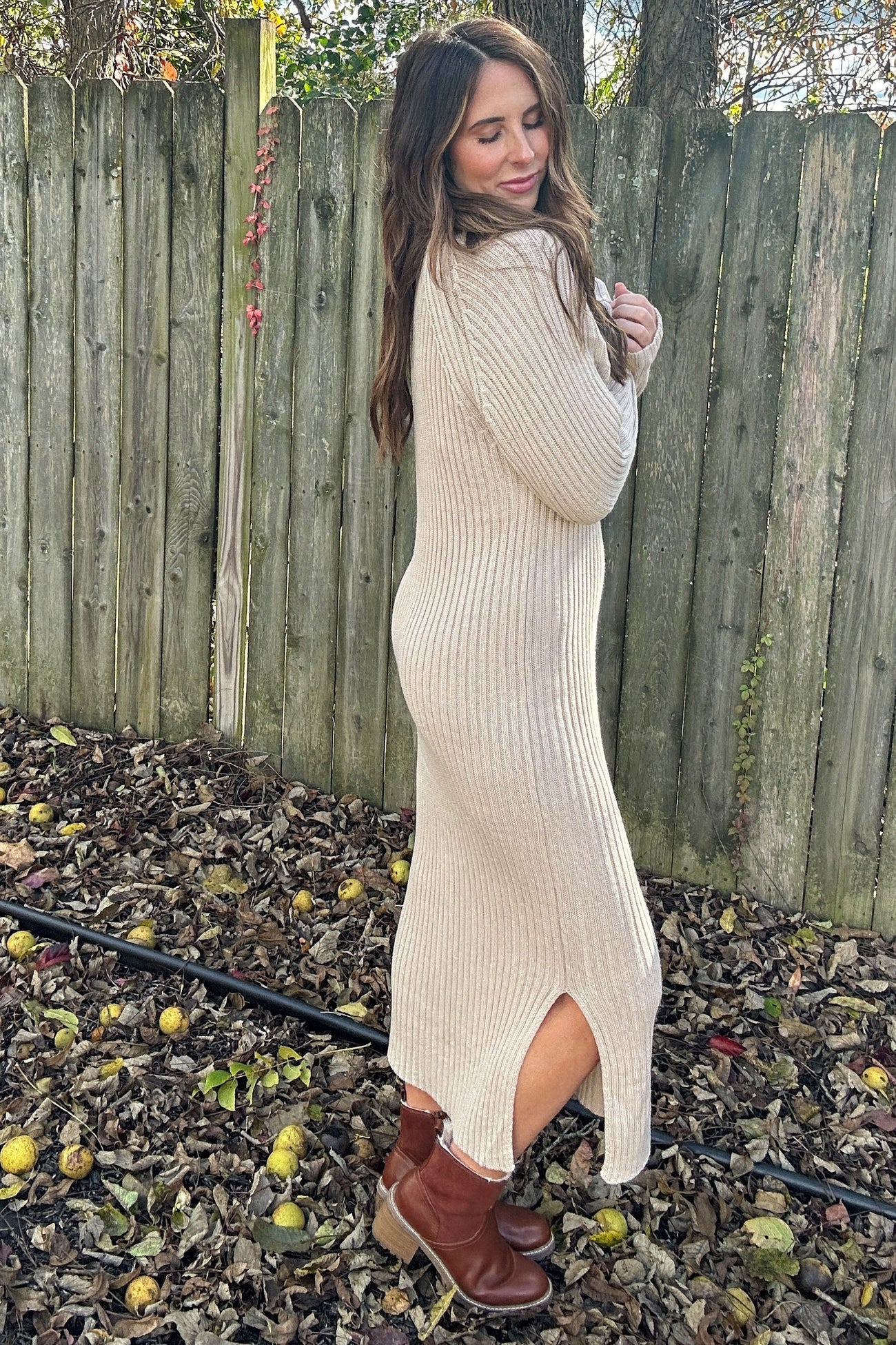 woman wearing a beige ribbed sweater dress with side slits and brown ankle boots, standing outdoors among fallen leaves