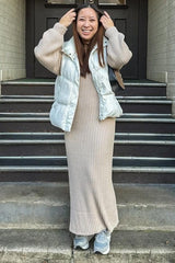 woman wearing a beige ribbed maxi dress and a white puffer vest, smiling on steps with gray sneakers