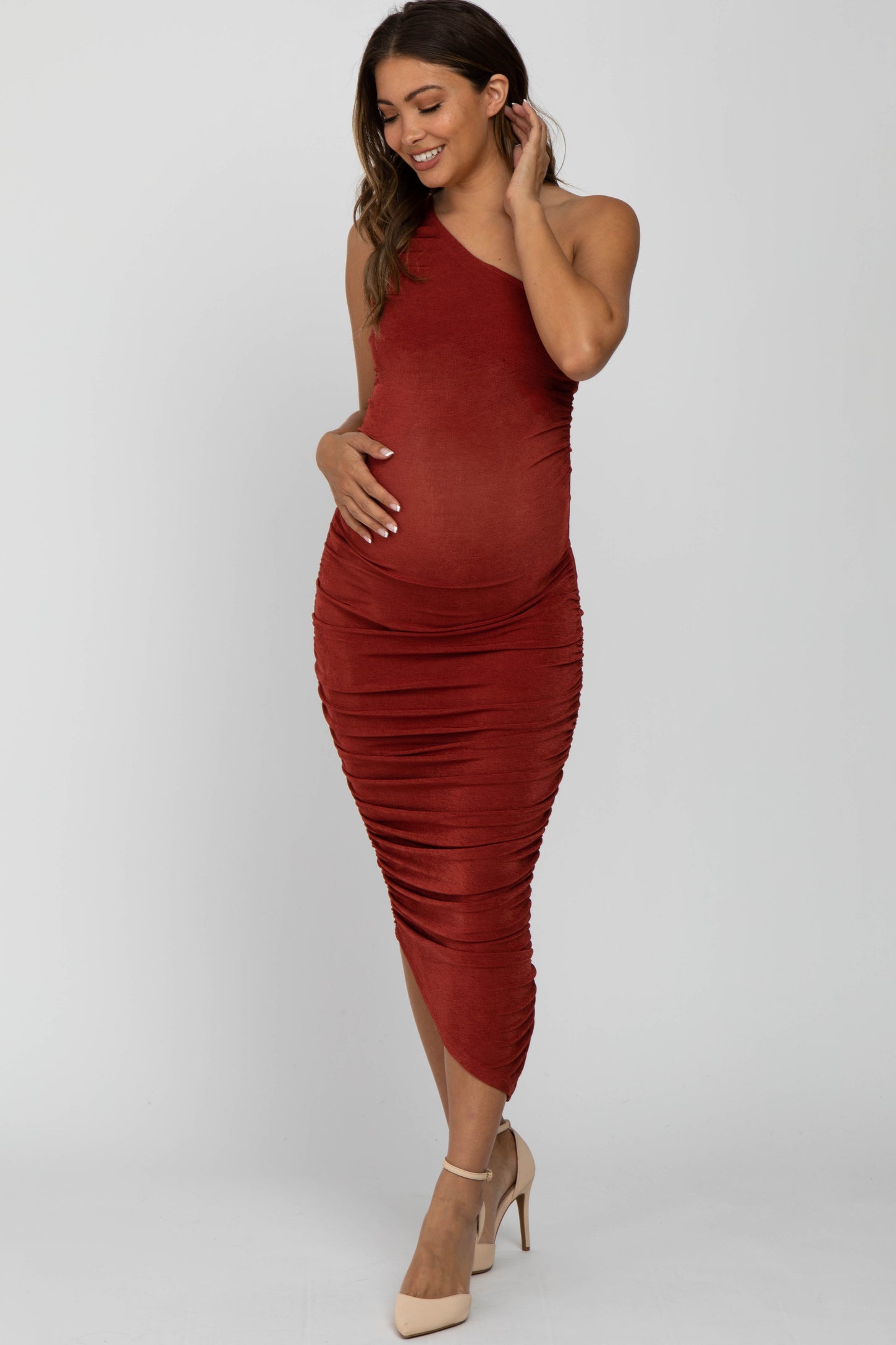 woman wearing a rust-colored one-shoulder ruched maternity dress and holding her baby bump