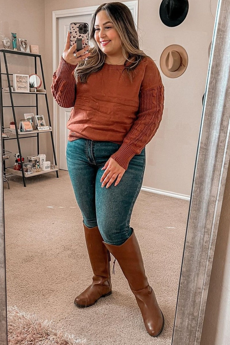 woman in rust orange cable knit sweater, blue jeans, and brown leather riding boots taking a mirror selfie