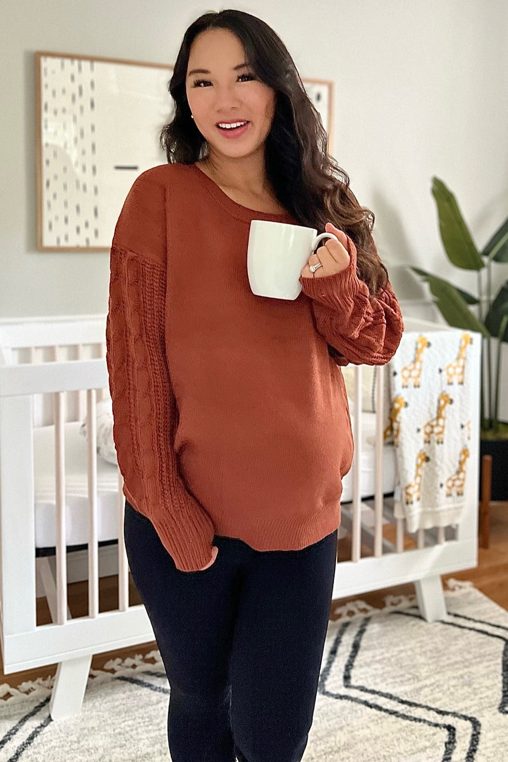 woman wearing a rust-colored maternity sweater with knitted sleeves and holding a white mug, showcasing her baby bump