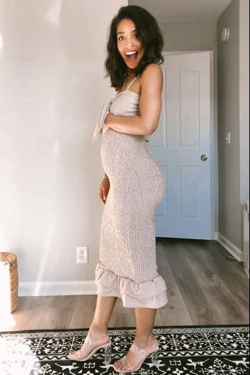 smiling pregnant woman in a beige smocked midi dress and clear heels standing in a room