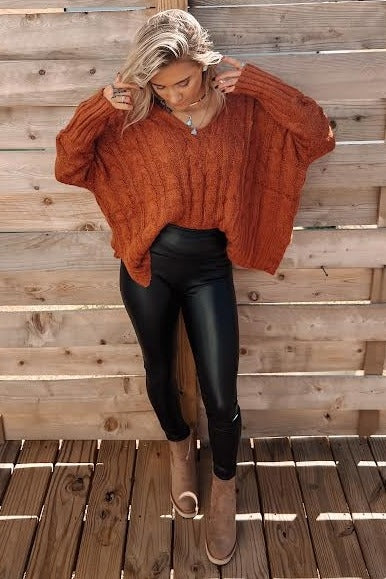 woman wearing an oversized rust-colored cable knit sweater and black leather leggings with taupe ankle boots