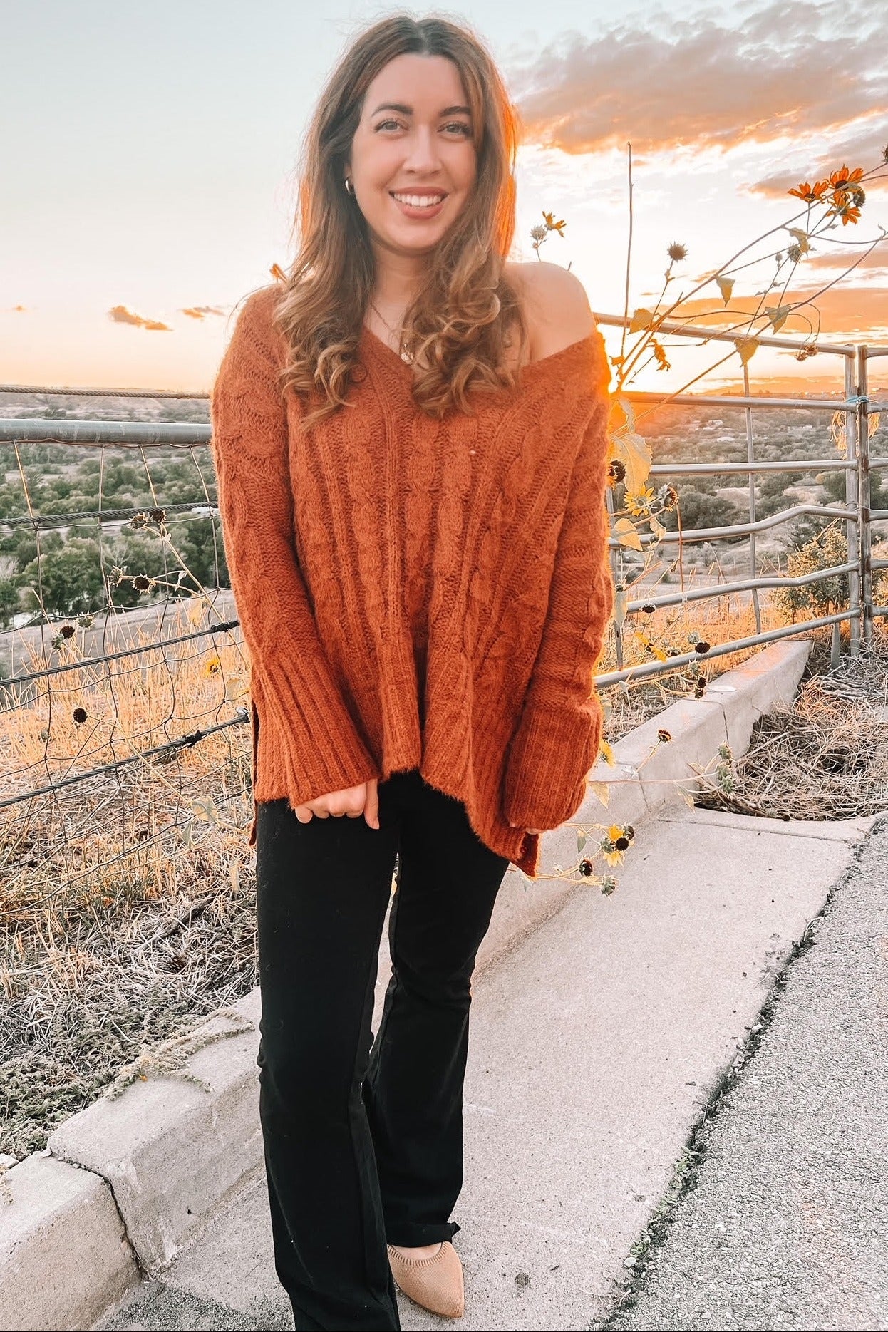 woman wearing an oversized rust-colored cable knit sweater and black flared pants, smiling outdoors at sunset