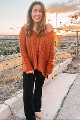woman wearing an oversized rust-colored cable knit sweater and black flared pants, smiling outdoors at sunset