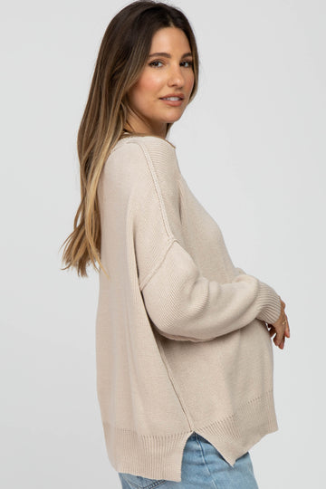 woman wearing a beige oversized maternity sweater with a side split and holding her baby bump