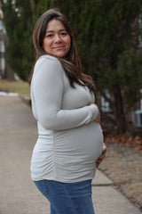 woman wearing a light grey long sleeve maternity top and holding her baby bump while standing outdoors