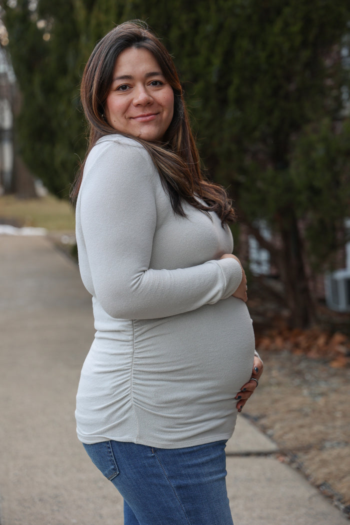 woman wearing a light grey long sleeve maternity top and holding her baby bump while standing outdoors