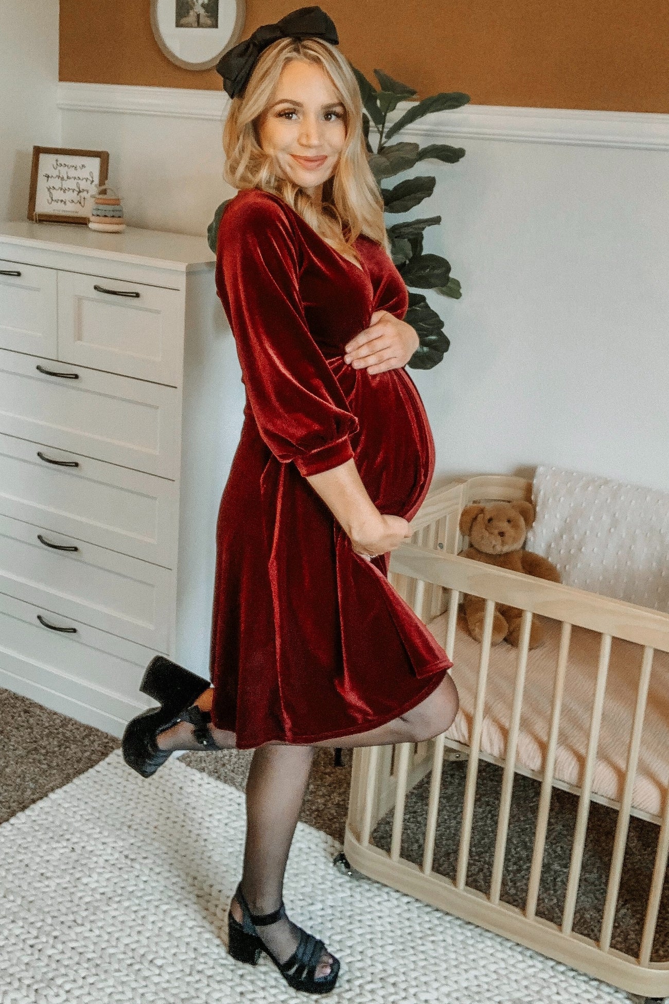 woman wearing a burgundy velvet maternity dress and holding her baby bump while standing next to a crib